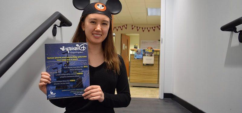 student wearing Mickey Mouse ears sits on a stair while holding a Disney internship flyer